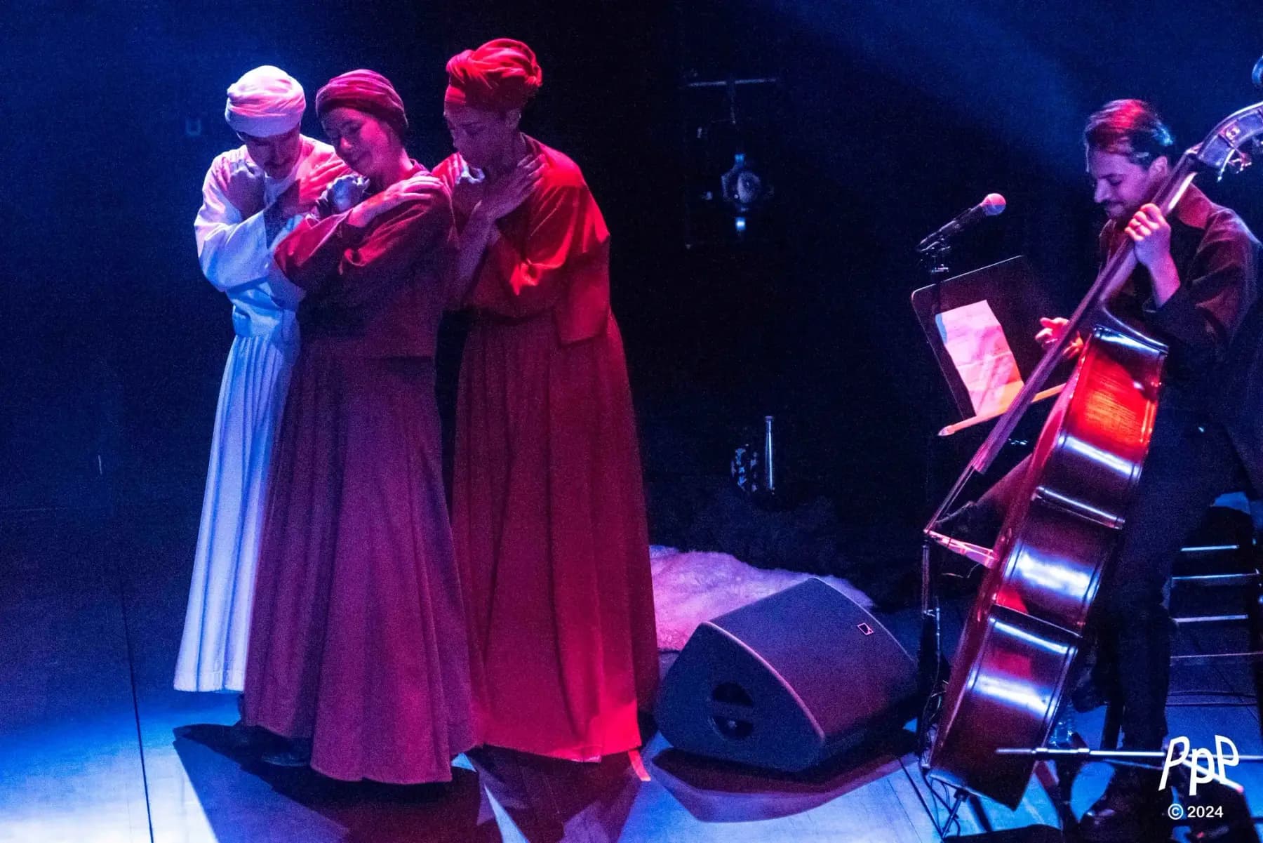Au bord de la scène, trois interprètes en longues robes et turbans réunis dans une même étreinte ; à côté, on joue de la contrebasse.