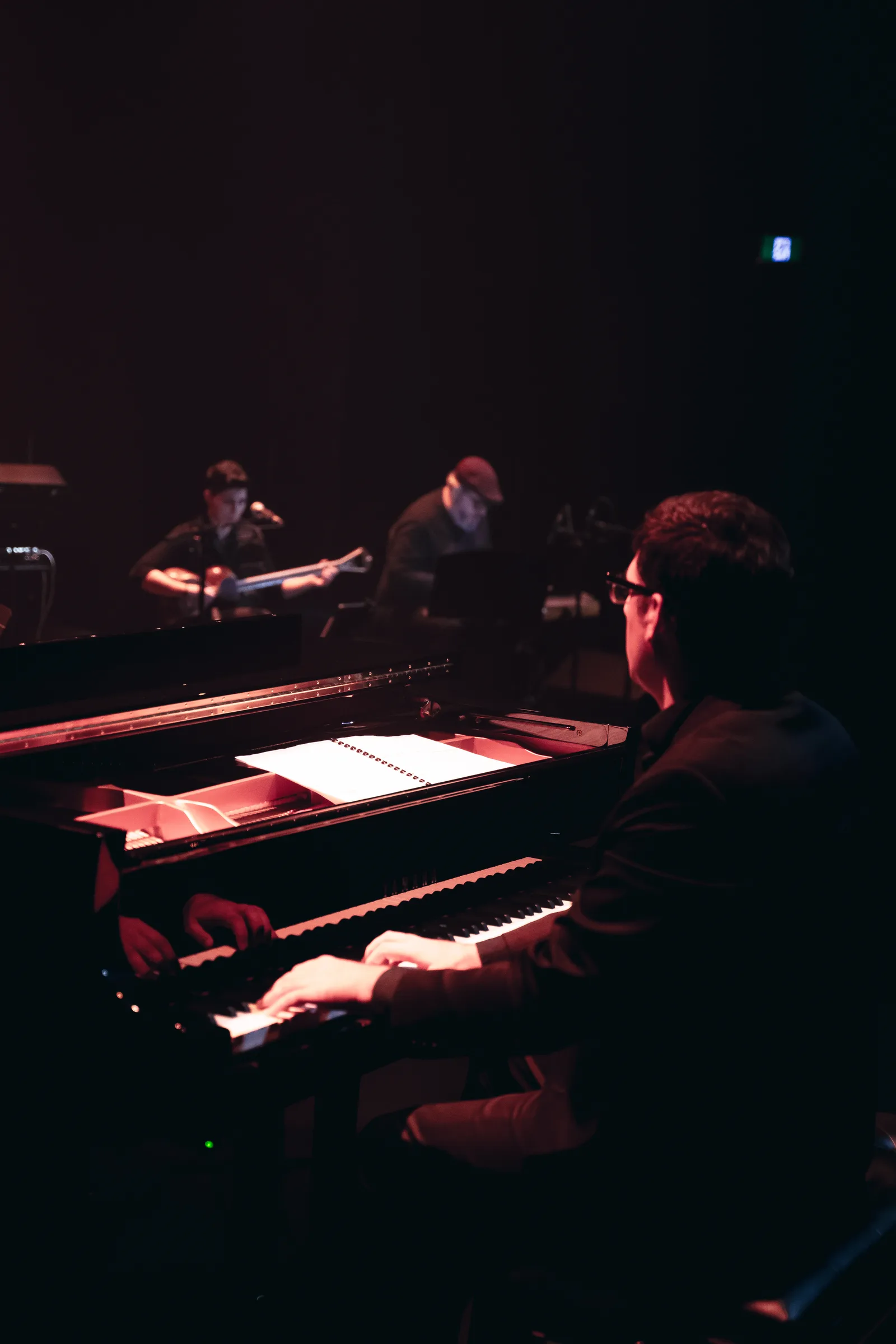 Au premier plan, le piano : les mains sur le clavier, la partition éclairée sur le pupitre. Au fond, le târ, sous la lumière de scène.